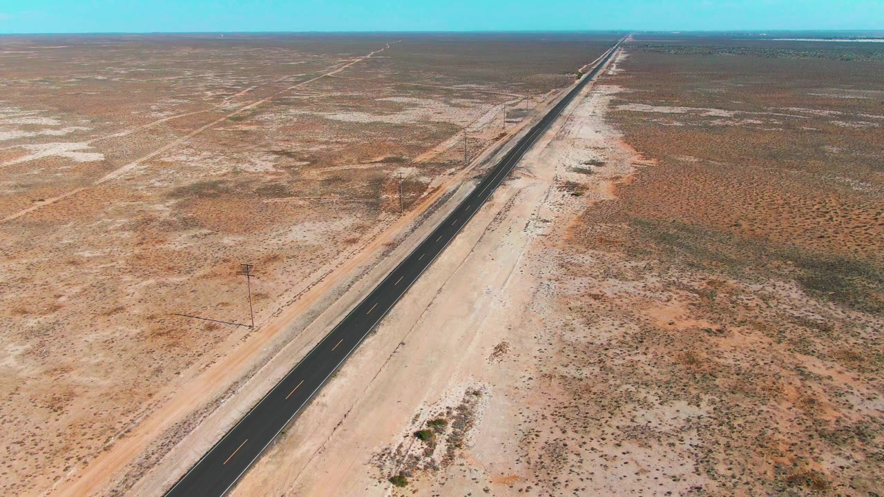 High oblique view over Guerrero Negro with a dark asphalt highway beside a broad pale sandy shoulder, low scrubland on both sides, and no visible people.