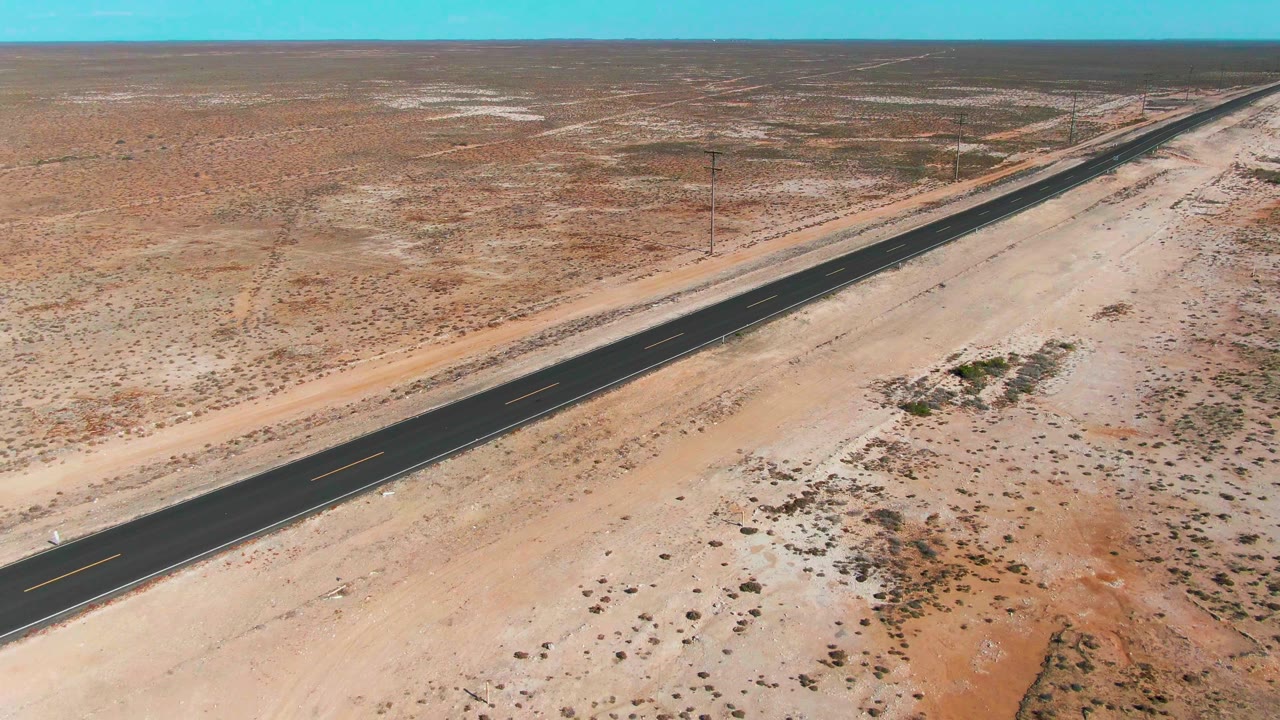 High aerial frame over Guerrero Negro desert shows the paved road stretching away across pale sand and low scrub, with utility poles beside it and no visible people.
