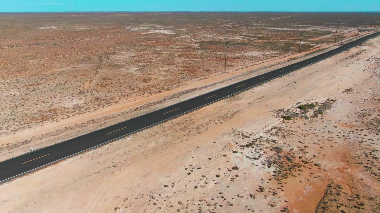 A drone-like view in Guerrero Negro frames the dark road at a diagonal, bordered by sandy tracks, scrub patches, and a broad blue sky band, with no visible people.