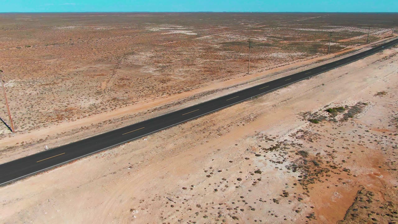 Aerial frame over Baja California Sur desert shows the highway running past utility poles and low vegetation across a flat sandy plain, with no visible people.