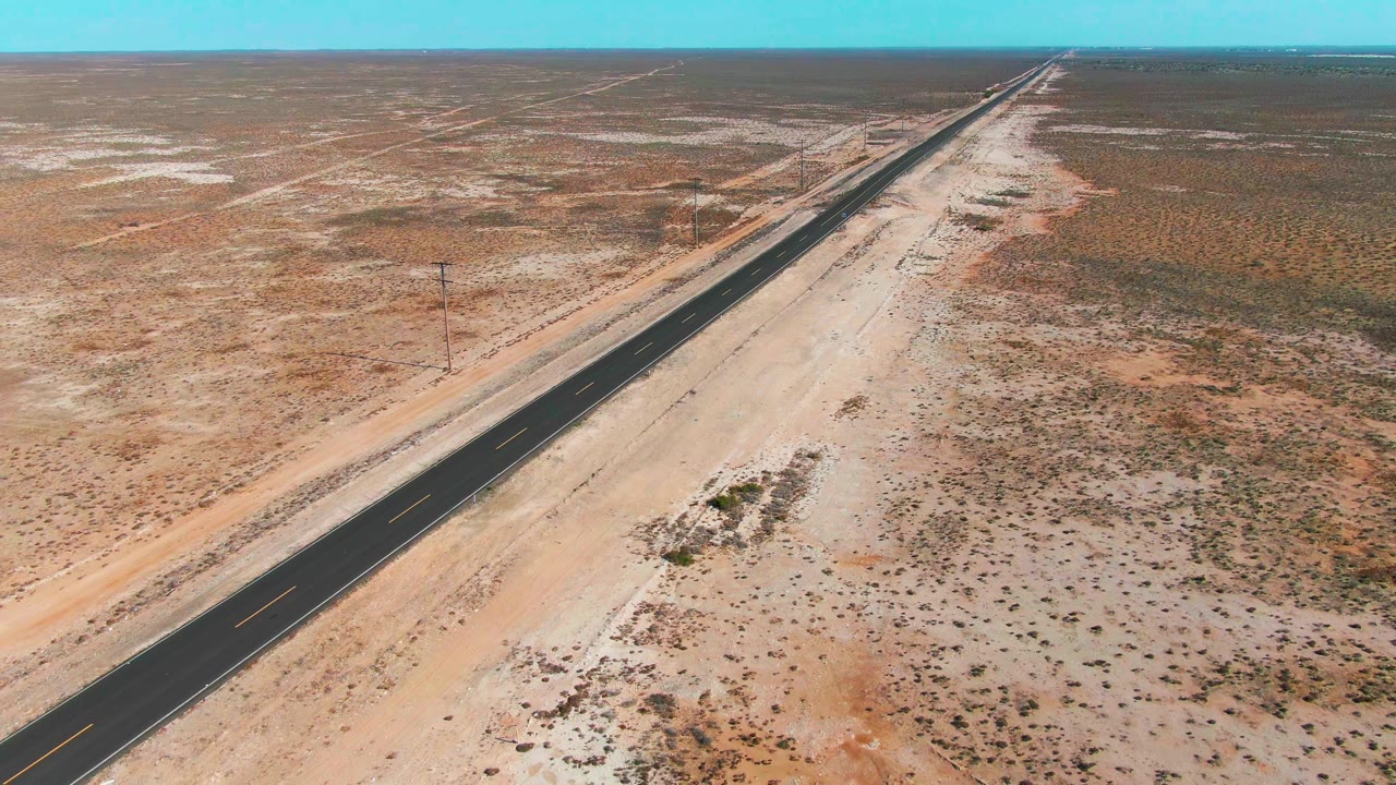 Aerial view near Guerrero Negro with the paved road running from the lower left toward the distant horizon, bordered by pale roadside sand, scattered scrub, and no visible people.