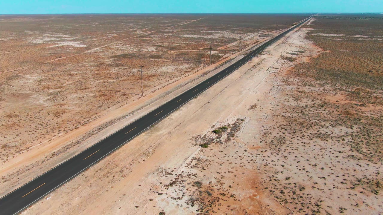 Drone-like aerial frame in Baja California Sur showing a dark straight road beside light sand berms and scrub patches, with a broad flat horizon and no visible people.