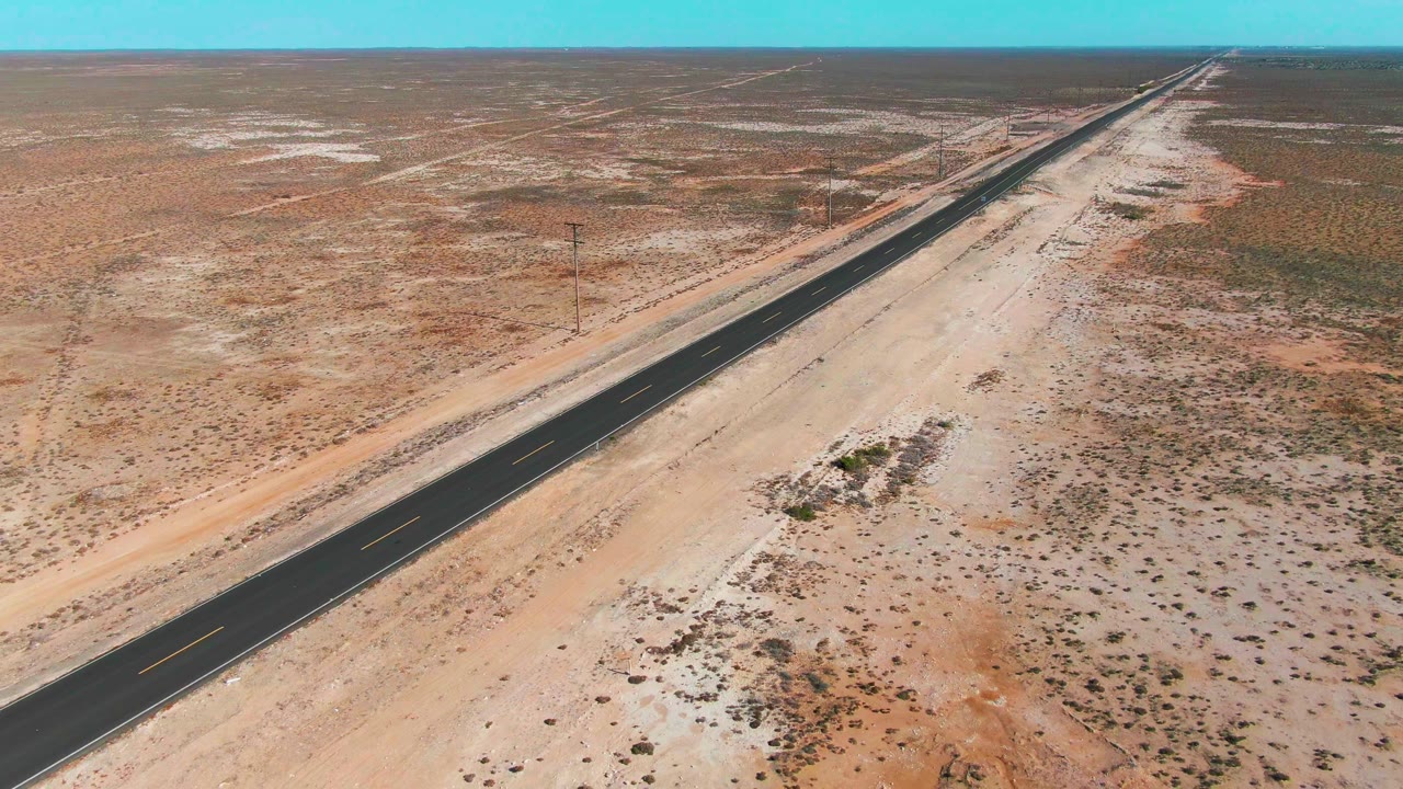 Aerial frame from Guerrero Negro showing the highway extending across scrubby desert, with utility poles along the roadside, pale sand patches, clear sky, and no visible people.