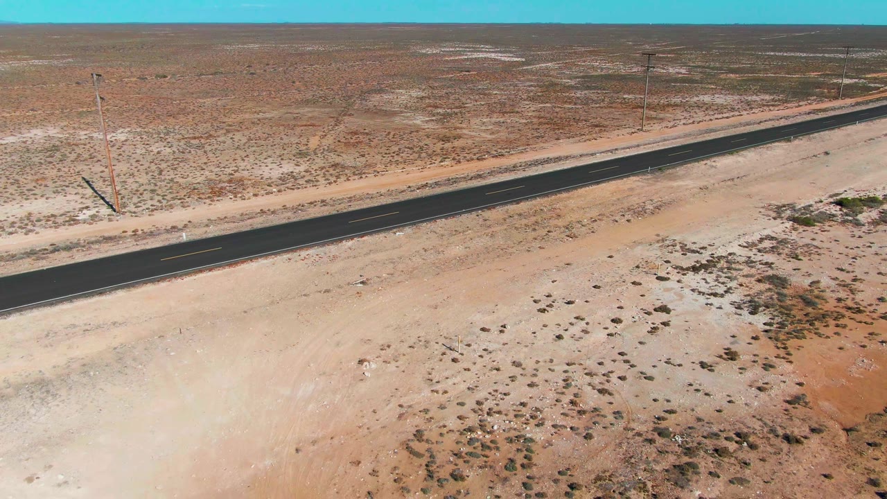 Aerial frame from Guerrero Negro, Baja California Sur showing a straight dark road across sandy desert, utility poles, clear blue sky, and no visible people.