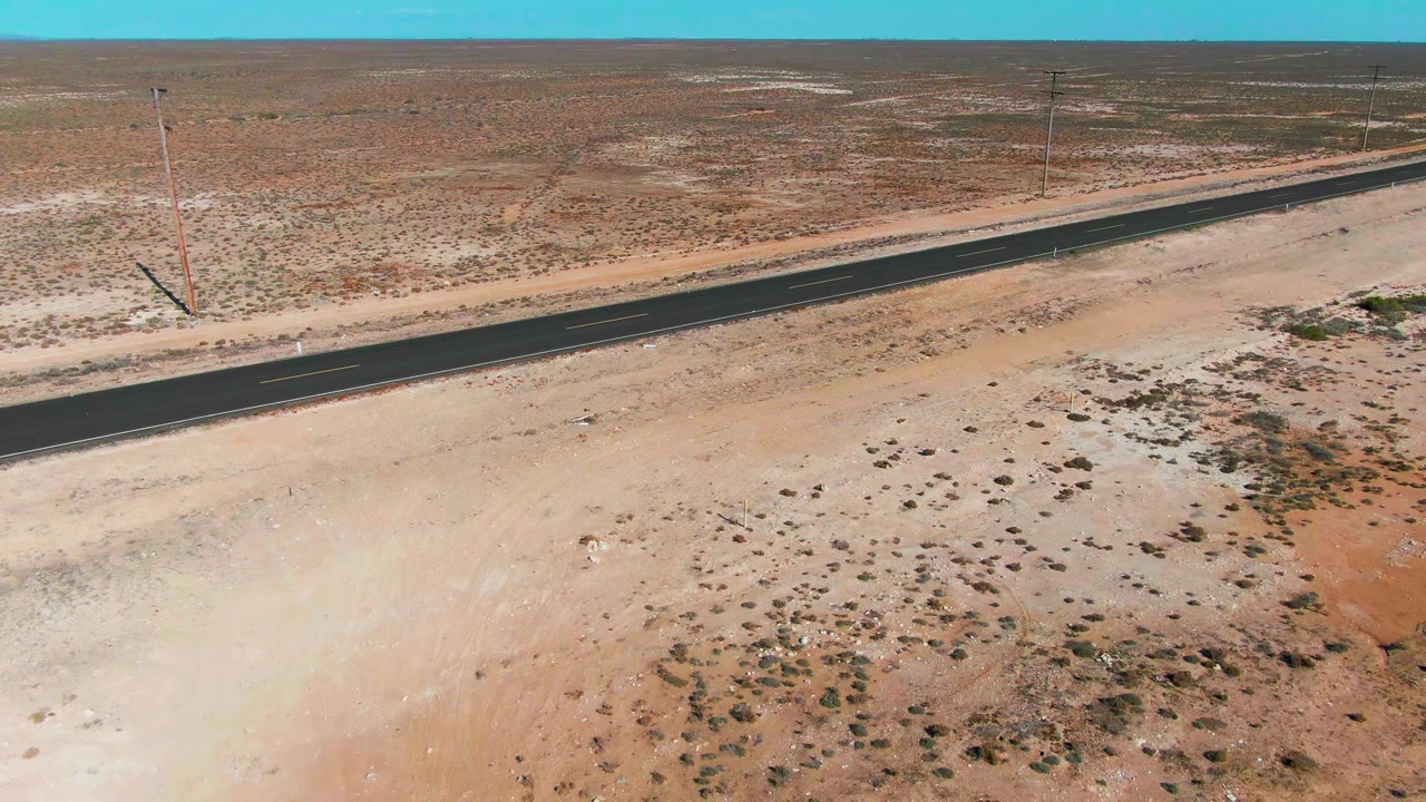 Straight paved road cutting through open sandy desert under blue sky.