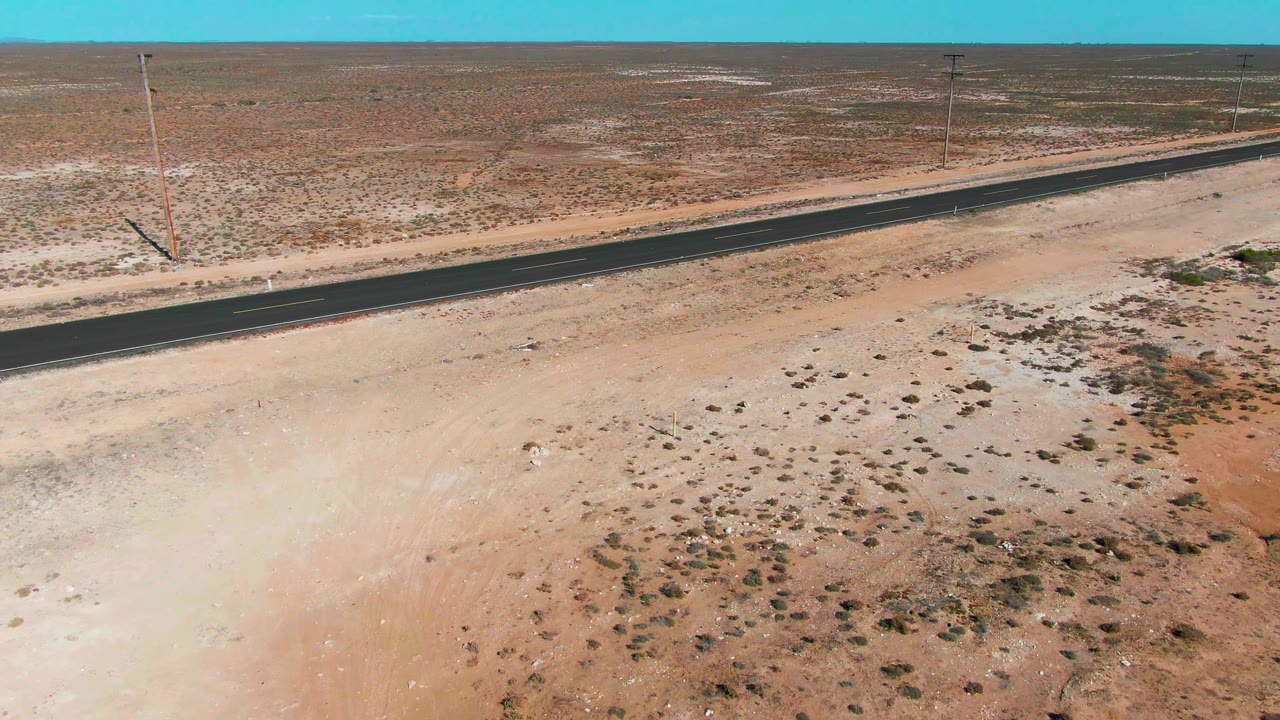 Aerial Guerrero Negro frame with the black road near the upper half of the image, pale sandy foreground, brown scrub patches, power poles, and no visible people.