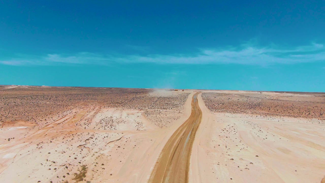 A high aerial view in Guerrero Negro, Baja California Sur centers the unpaved road as it runs through sandy scrubland toward a pale dust cloud, with no visible people.