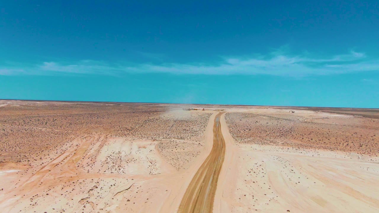 Aerial desert landscape with a dirt road entering from the lower frame and dust ahead.