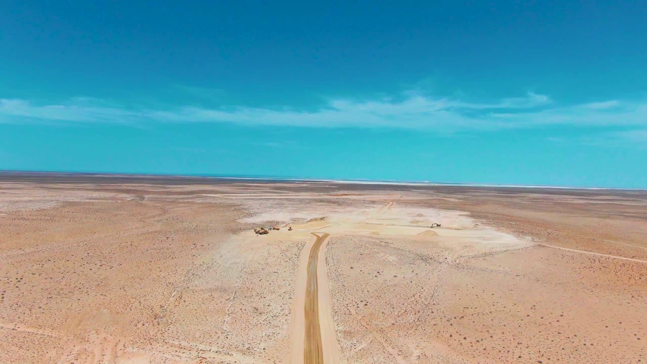 Aerial frame from Guerrero Negro, Baja California Sur showing a dirt road through sandy desert, a small vehicle near the pale flat area, blue sky, and no visible people.