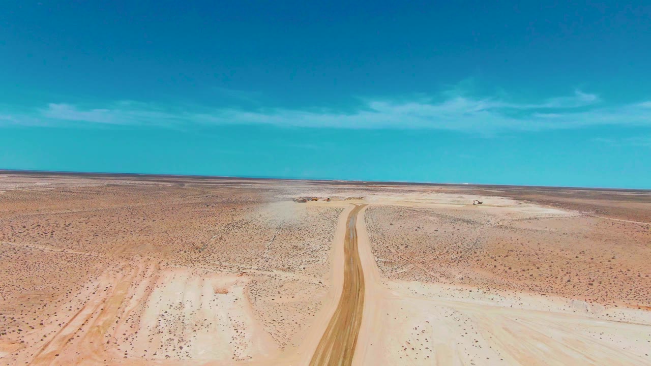 Opening aerial frame over Guerrero Negro desert terrain, with a pale dirt road running toward a flat horizon, a tiny vehicle near the road, thin clouds above, and no visible people.