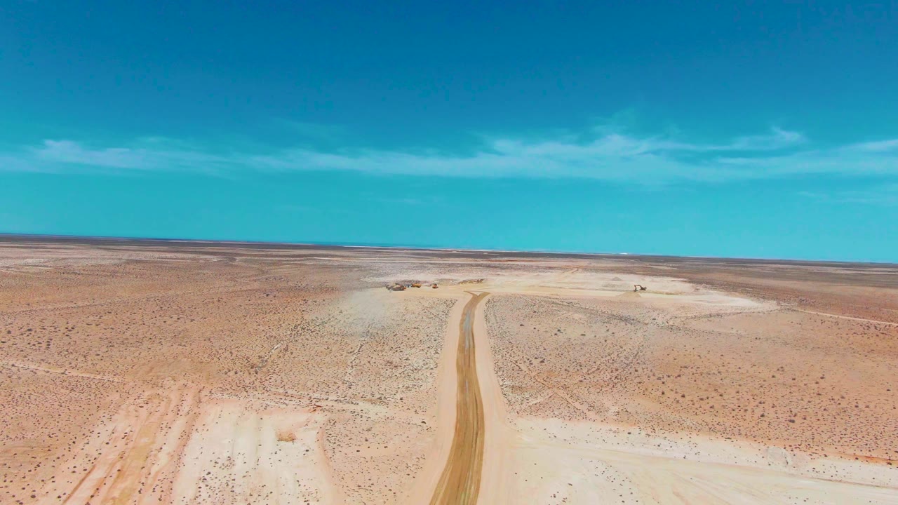 Guerrero Negro aerial frame with the dirt road rising from the lower edge toward the horizon, sparse desert vegetation on both sides, a small vehicle near the upper road area, and no visible people.