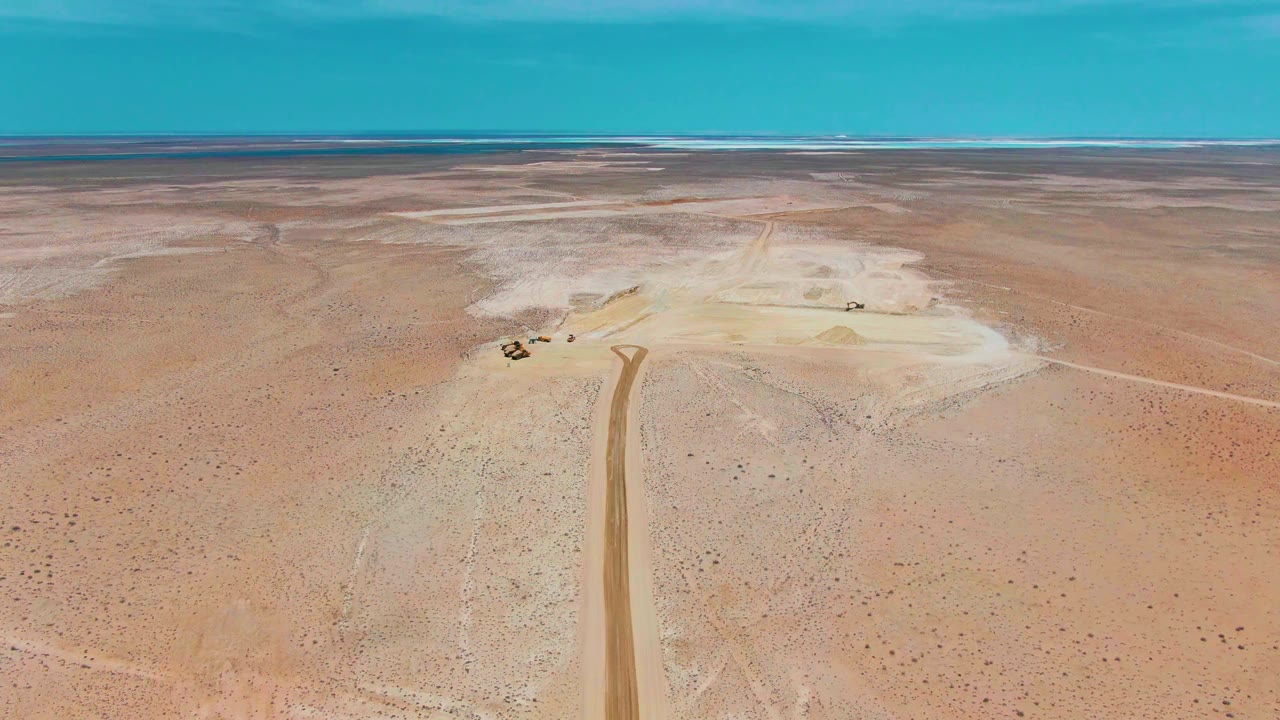 Wide aerial view in Guerrero Negro, Baja California Sur, showing a dirt road crossing sandy desert toward small construction vehicles, with salt flat tones and no visible people.