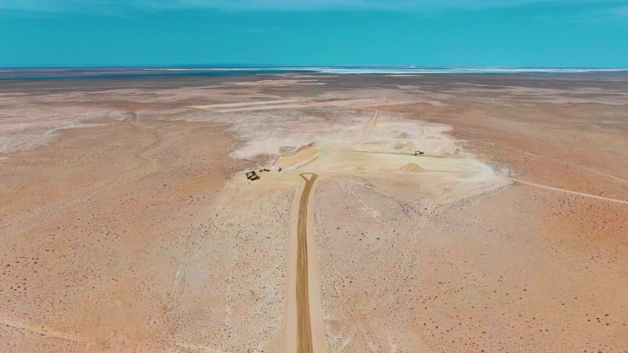 Aerial frame over Guerrero Negro desert with a straight dirt road entering from the bottom edge, small machinery near pale ground, distant dark waterline, and no visible people.
