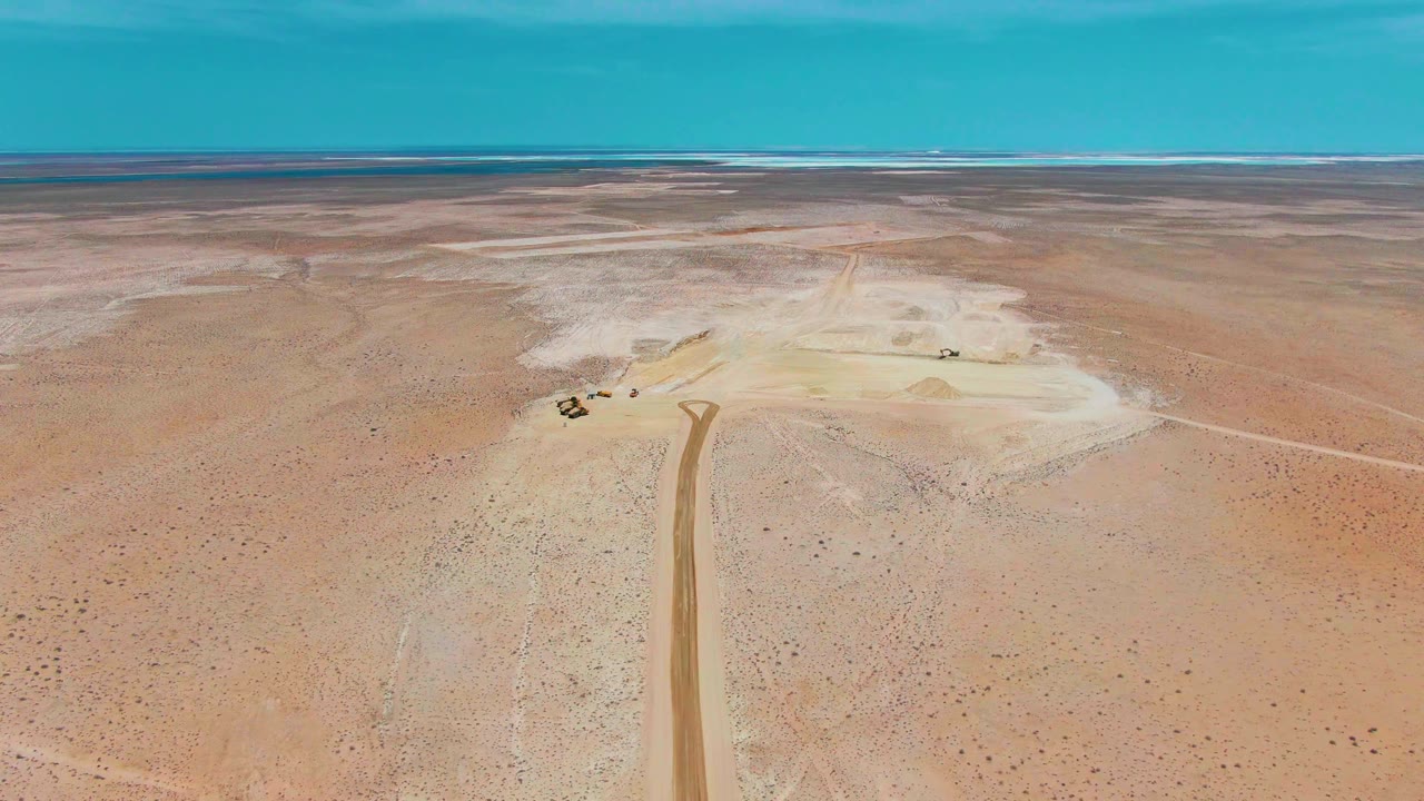 High drone view in Guerrero Negro showing the road aligned near center, small vehicles beside a light-toned work area, open sandy terrain, blue sky, and no visible people.