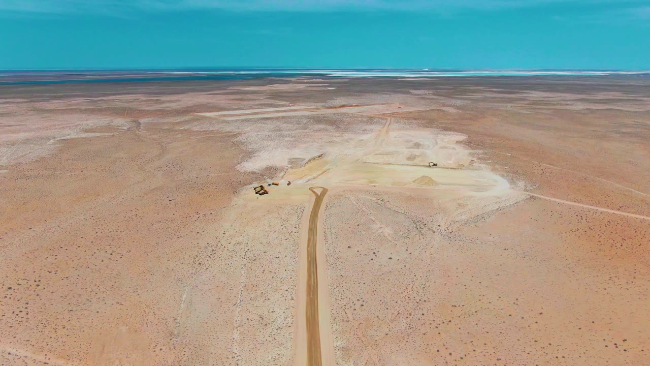 Aerial desert landscape with dirt road, small vehicles, and distant flat horizon.