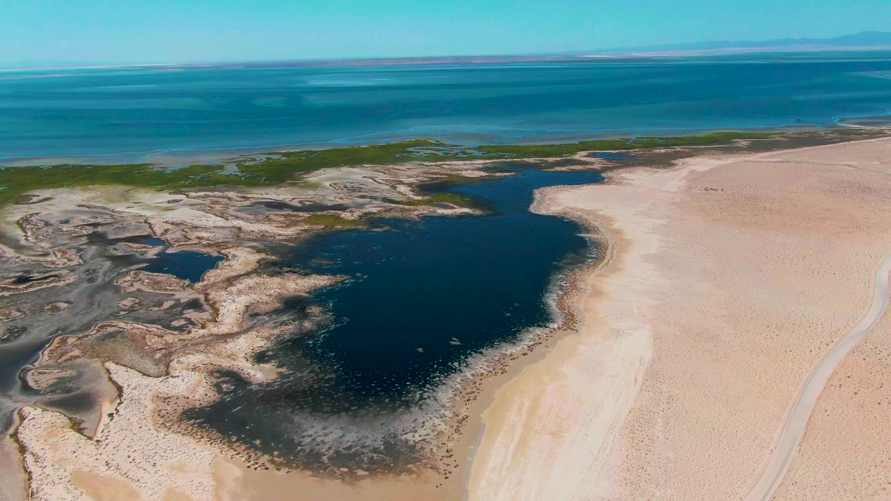 Aerial view in Guerrero Negro, Baja California Sur with the dark lagoon curving along the sandy beach, mudflat channels at left, open blue water beyond, and no visible people.