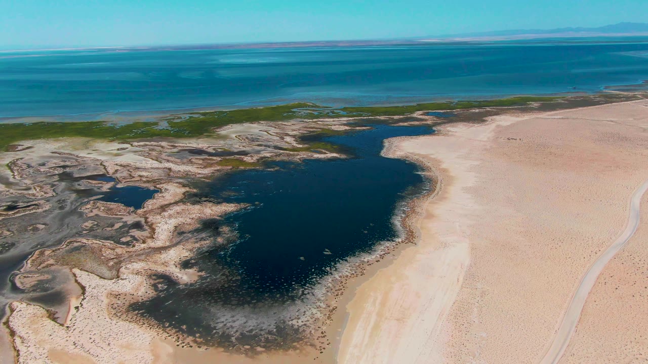 High aerial frame over Guerrero Negro, Baja California Sur showing dark lagoon water centered between pale sand and patterned mudflats, with ocean and no visible people in the distance.