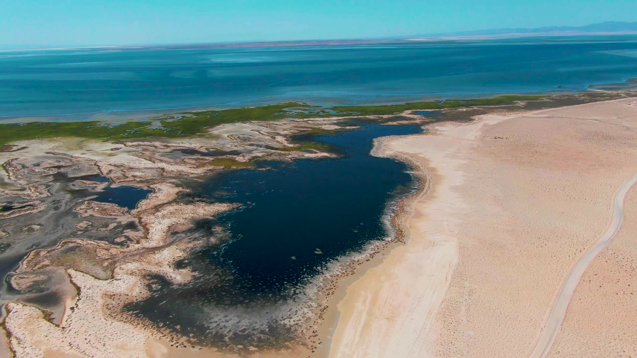 Aerial frame from Guerrero Negro, Baja California Sur with the lagoon slightly lower in view, empty sand to the right, mudflat texture to the left, blue ocean above, and no visible people.