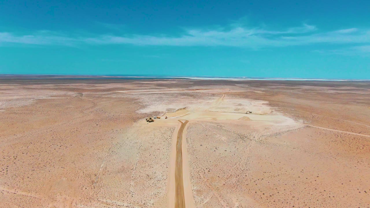 Aerial view in Guerrero Negro, Baja California Sur, with a dirt road entering from the bottom of the frame toward pale desert flats, small vehicles or equipment nearby, a distant horizon, and no visible people.