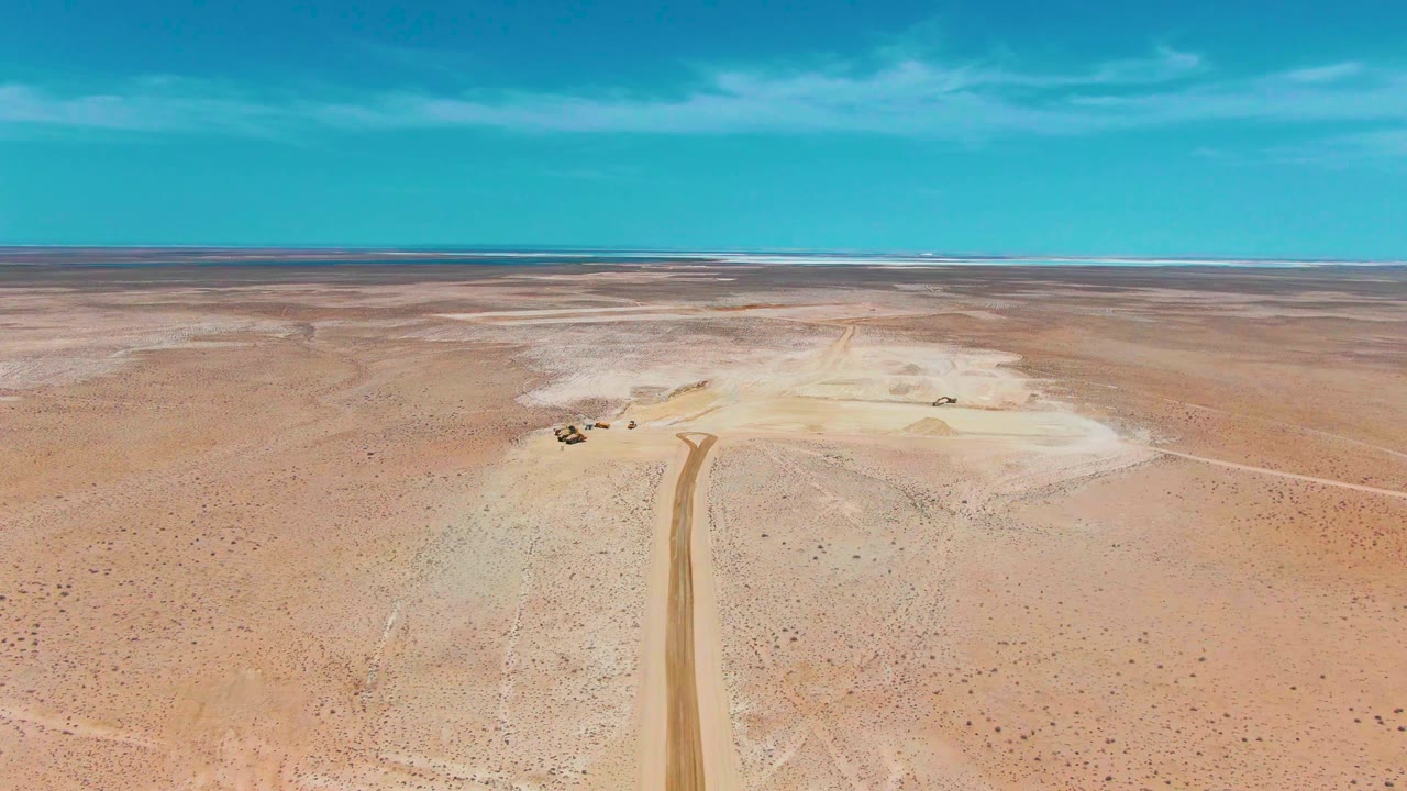 Aerial view of the Guerrero Negro desert with a dirt road cutting through sparse scrub, pale sandy areas on both sides, small vehicle shapes near the track, a low distant horizon, and no visible people.