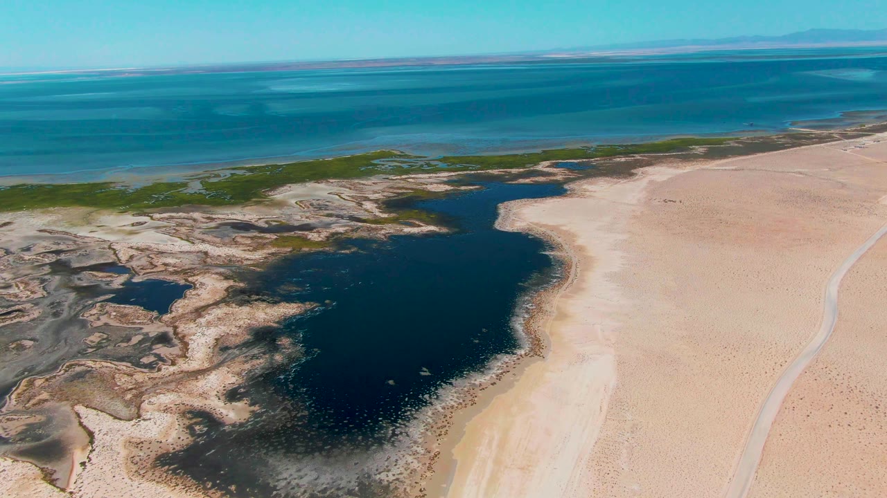 Aerial frame over Guerrero Negro with the lagoon centered, rough wetland edges at left, pale sand flats at right, blue ocean in the background, and no visible people.