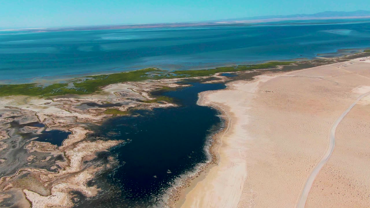 Wide aerial frame in Guerrero Negro showing the lagoon slightly more centered, with dry sand flats on the right, green shoreline patches on the left, ocean bands, and no visible people.