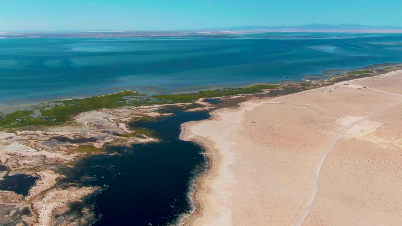 High aerial frame from Guerrero Negro showing turquoise ocean, a broad pale beach, dark irregular shallow water along the shore, green vegetation patches, and no visible people.