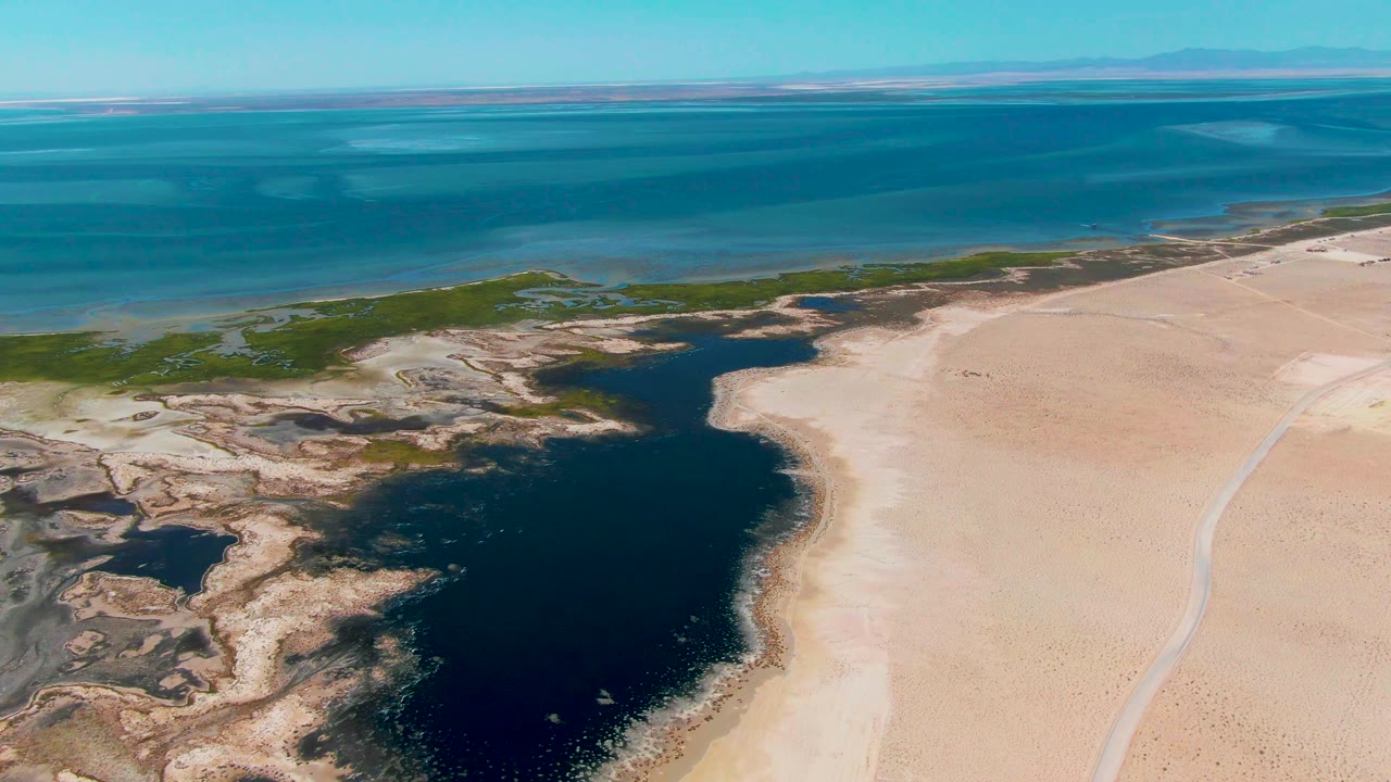 Aerial view in Guerrero Negro, Baja California Sur, with the lagoon centered lower in frame, sand flats to the right, shallow green shoreline areas to the left, and no visible people.