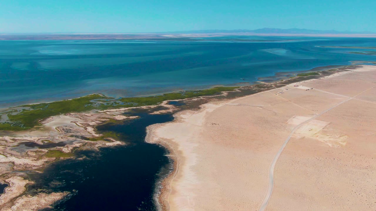 At 0:00, an aerial view over Guerrero Negro shows blue coastal water, pale sand, green marsh edges, a dark lagoon in the lower left, distant low land, and no visible people.