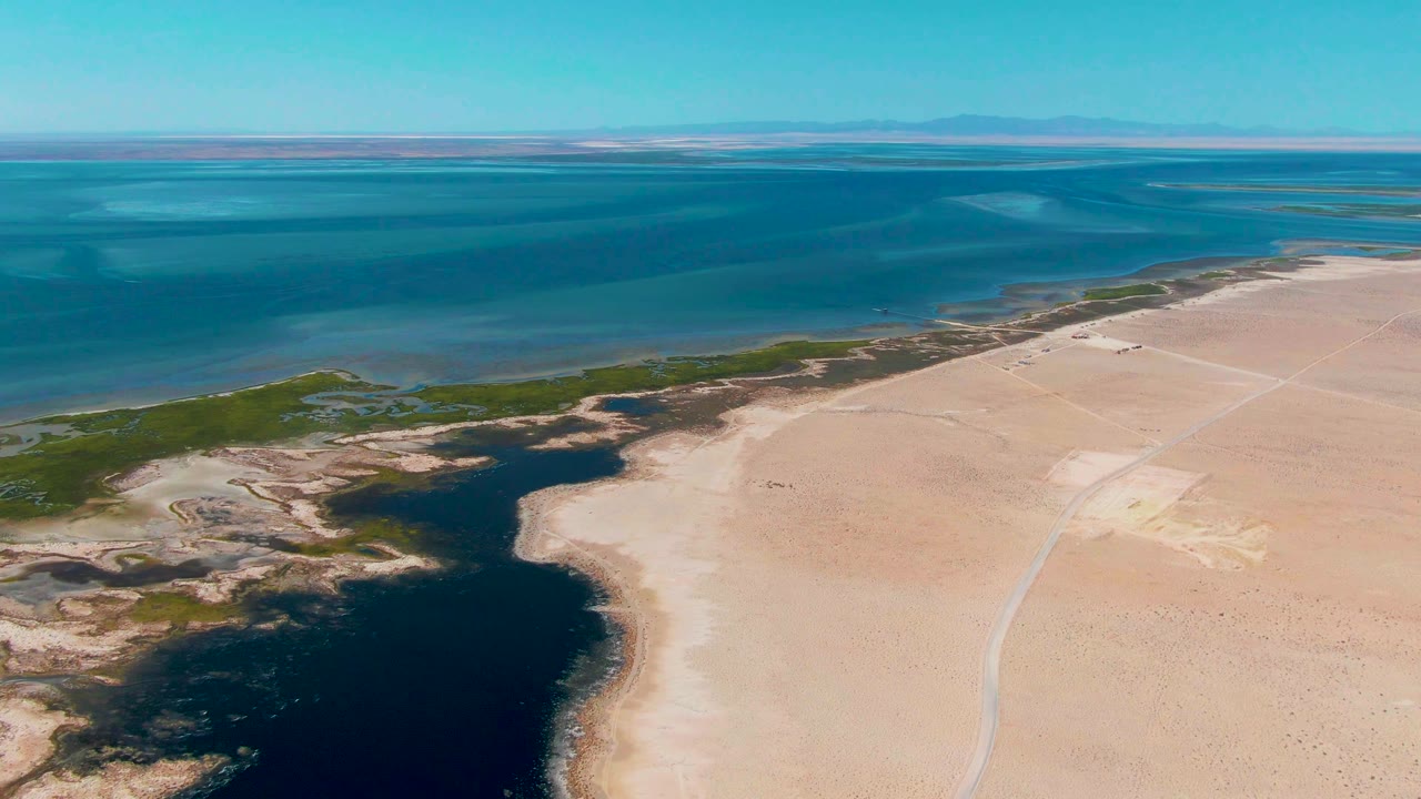 At 0:02, the aerial frame shows Guerrero Negro blue water meeting a pale sandy shoreline, with irregular green wetland patches and a dark inlet near the lower left, and no visible people.
