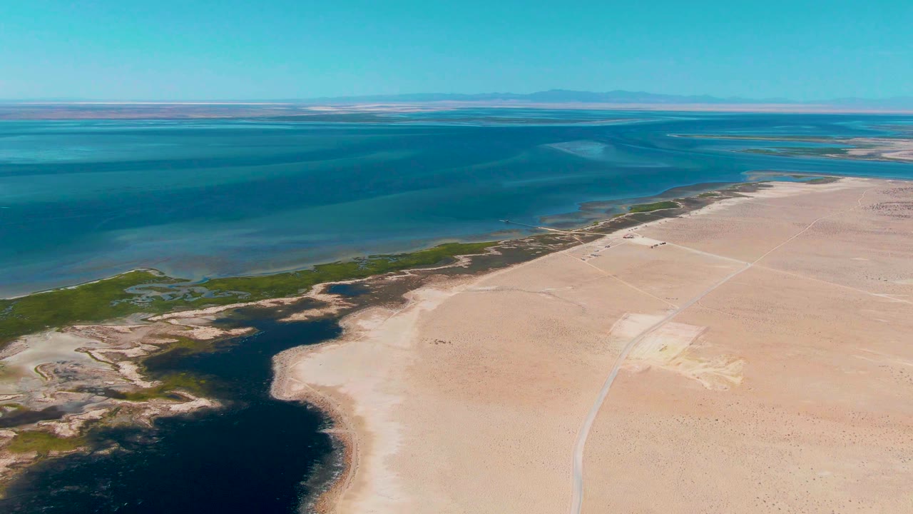 Opening aerial frame over Guerrero Negro, Baja California Sur, with dark lagoon water on the left, pale sandy land on the right, green shoreline vegetation, clear sky, and no visible people.