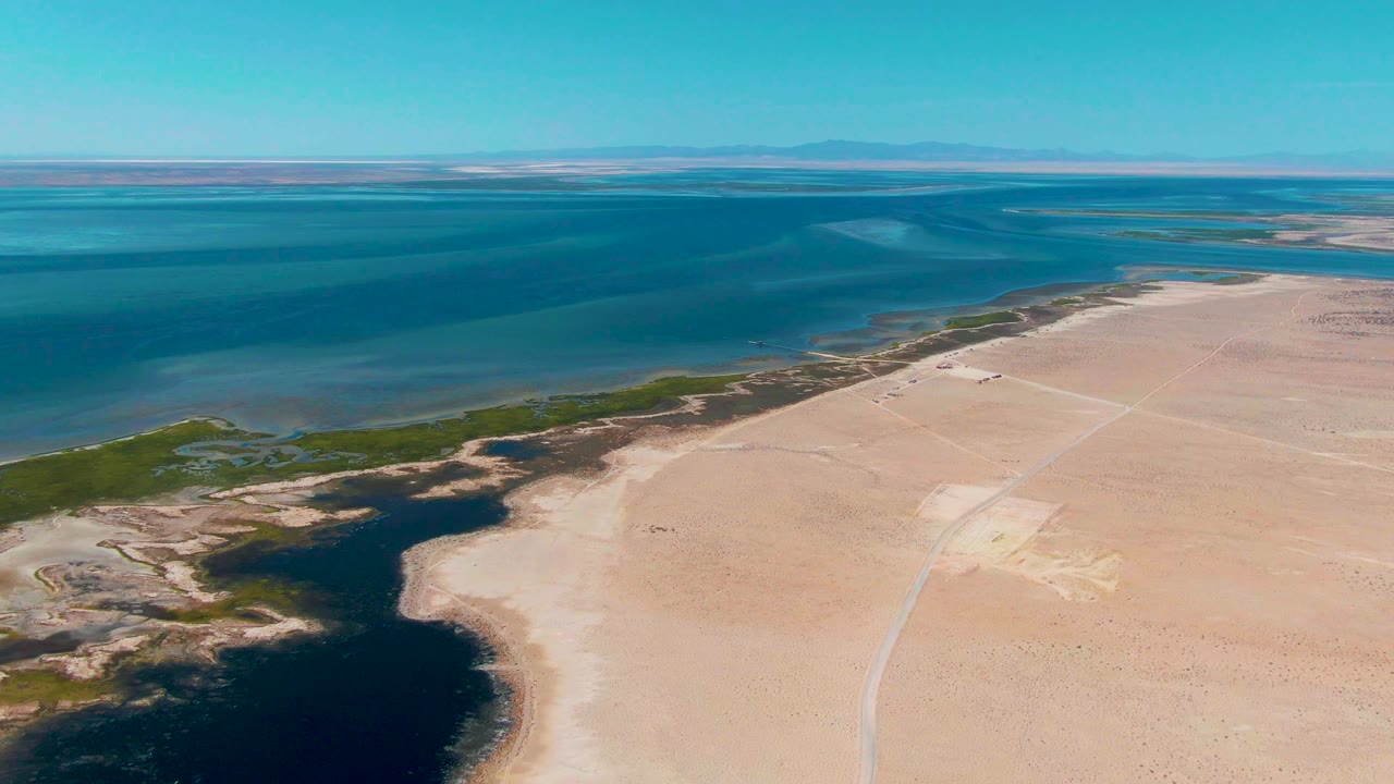 Aerial frame over Guerrero Negro showing broad blue lagoon water across the upper left, exposed sand on the right, a green wetland strip along the shore, distant flats, and no visible people.