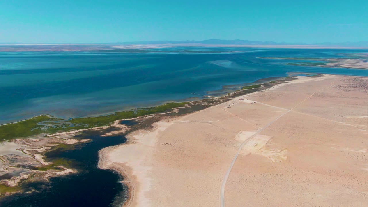 Later aerial frame over the Guerrero Negro shoreline, with a dark water pocket at lower left, pale dry land across the right side, green marsh-like edges, clear blue sky, and no visible people.