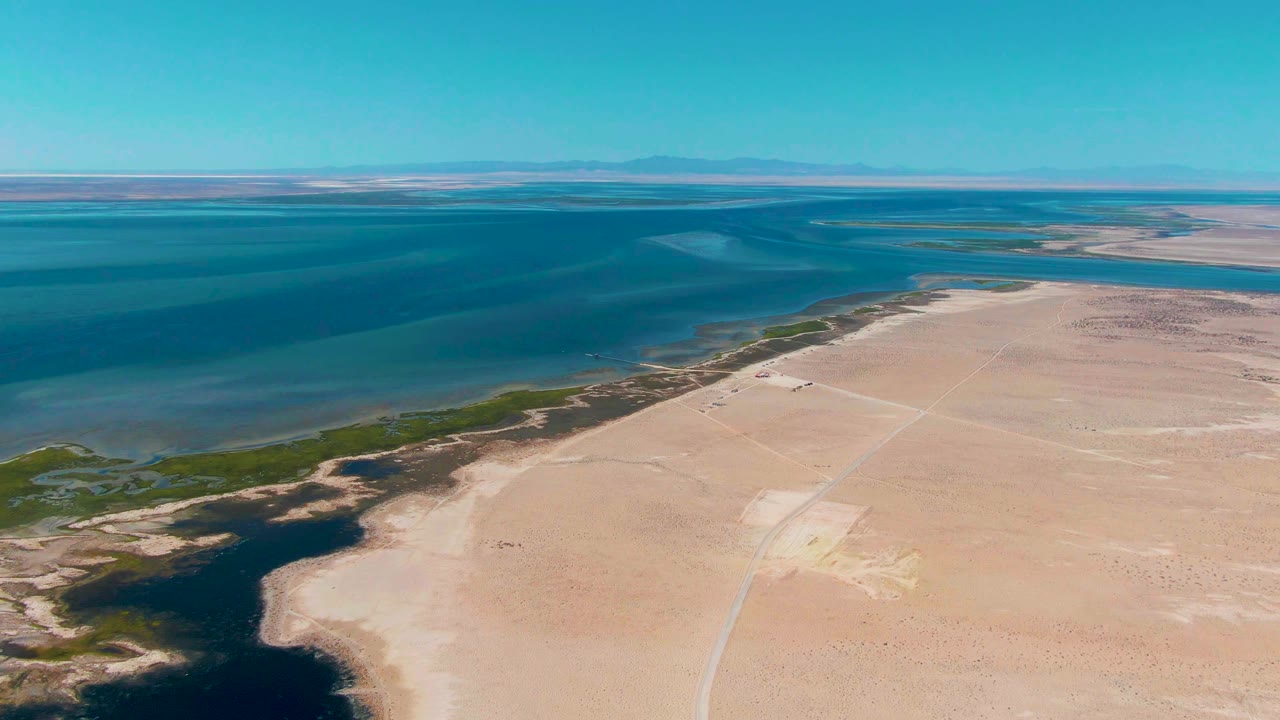 Guerrero Negro aerial frame with a darker inlet shape near the lower left, green vegetation marking the shore, open blue water beyond, pale sand flats, and no visible people.