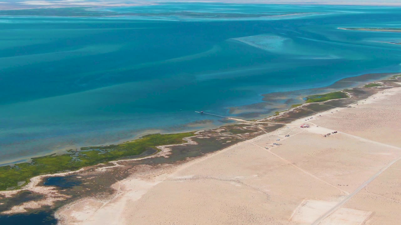 At one second, the Guerrero Negro coast remains framed from above with blue water across the top, sandy terrain below, vegetation along the shore, and no visible people.