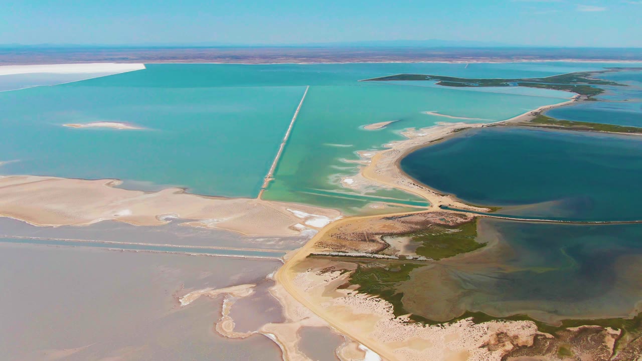 Second aerial frame from Guerrero Negro showing blue-green salt ponds, a narrow causeway, pale evaporation flats, darker water to the right, and no visible people.