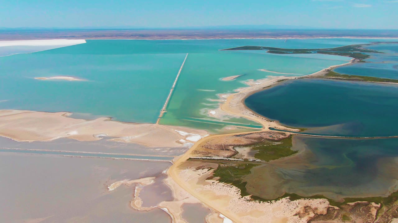 Aerial frame over Guerrero Negro with turquoise evaporation ponds, white salt deposits, a curved sandy edge, dark lagoon water, distant channels, and no visible people.