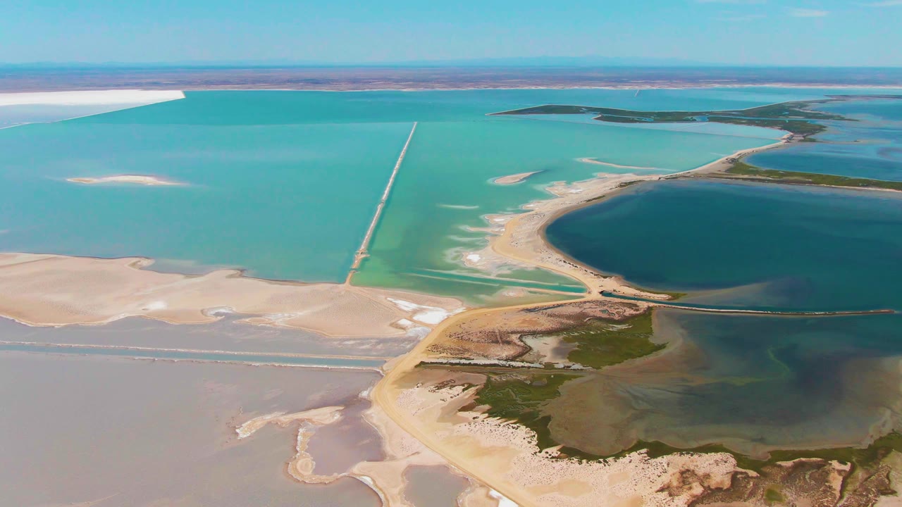 Mid-clip aerial frame in Guerrero Negro showing bright blue-green evaporation ponds, broad pale salt flats in the foreground, the straight causeway, and no visible people.