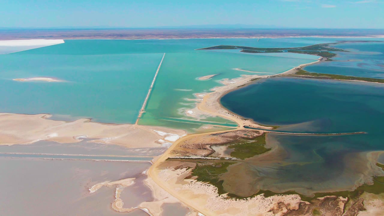 Wide aerial frame over Guerrero Negro salt lake with turquoise shallows, a dark blue lagoon edge, pale sandy flats, a straight causeway or pipeline, and no visible people.