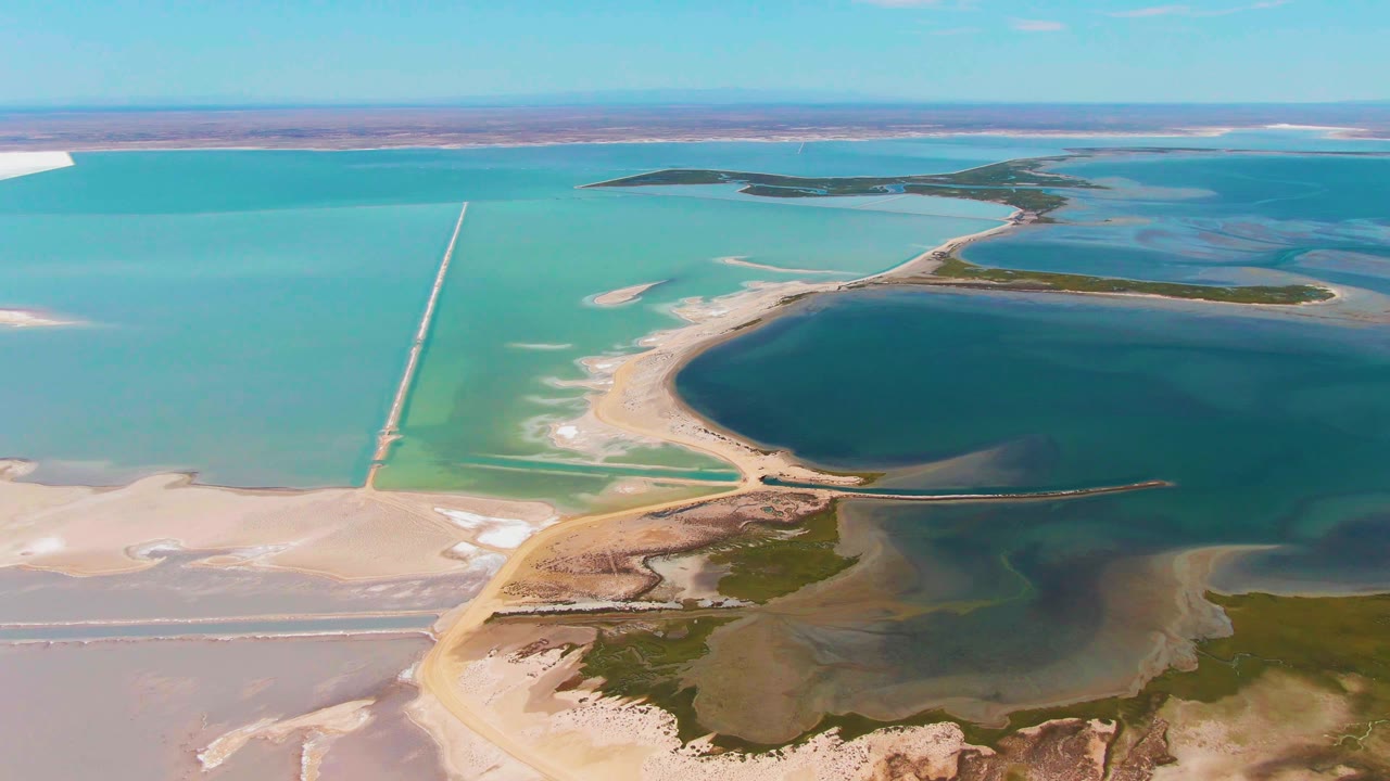 Aerial view in Guerrero Negro with the long causeway more centered, pale salt flats at lower left, dark pond water in the middle, distant turquoise basins, and no visible people.