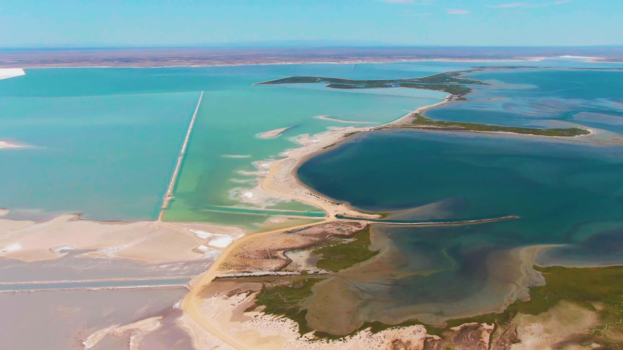 Guerrero Negro aerial frame showing blue-green evaporation ponds, a narrow road or causeway crossing the water, white mineral edges, sandy flats, vegetation patches, and no visible people.