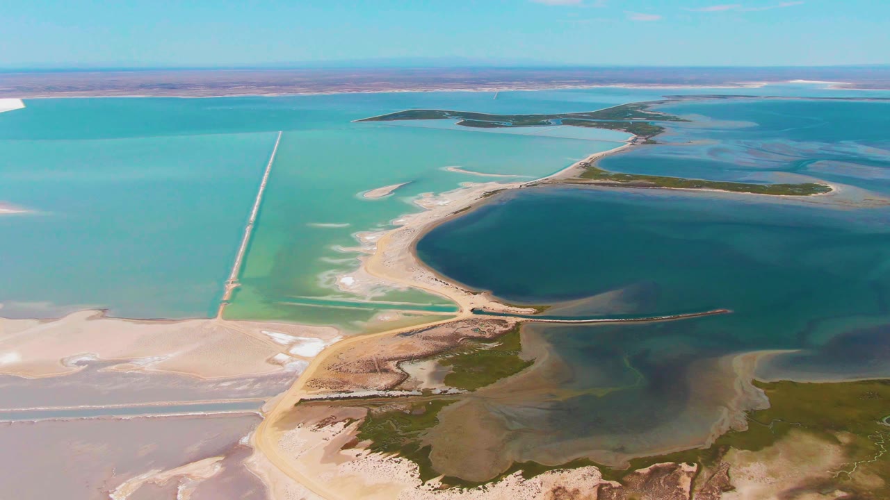 Later aerial frame over Guerrero Negro salt ponds with a dark central basin, pale salt flats on the left, turquoise water beyond, causeway lines, green marsh-like patches, and no visible people.