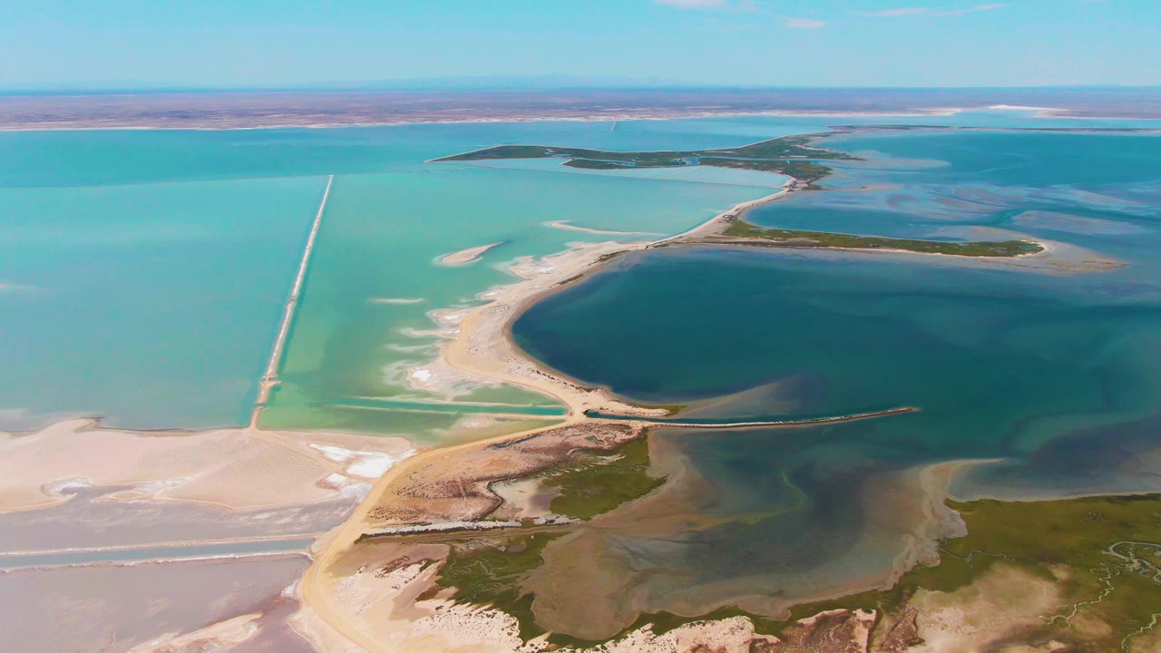 Aerial view over Guerrero Negro evaporation ponds with turquoise water, a dark blue basin, pale salt flats, straight causeways, distant flat land, and no visible people.