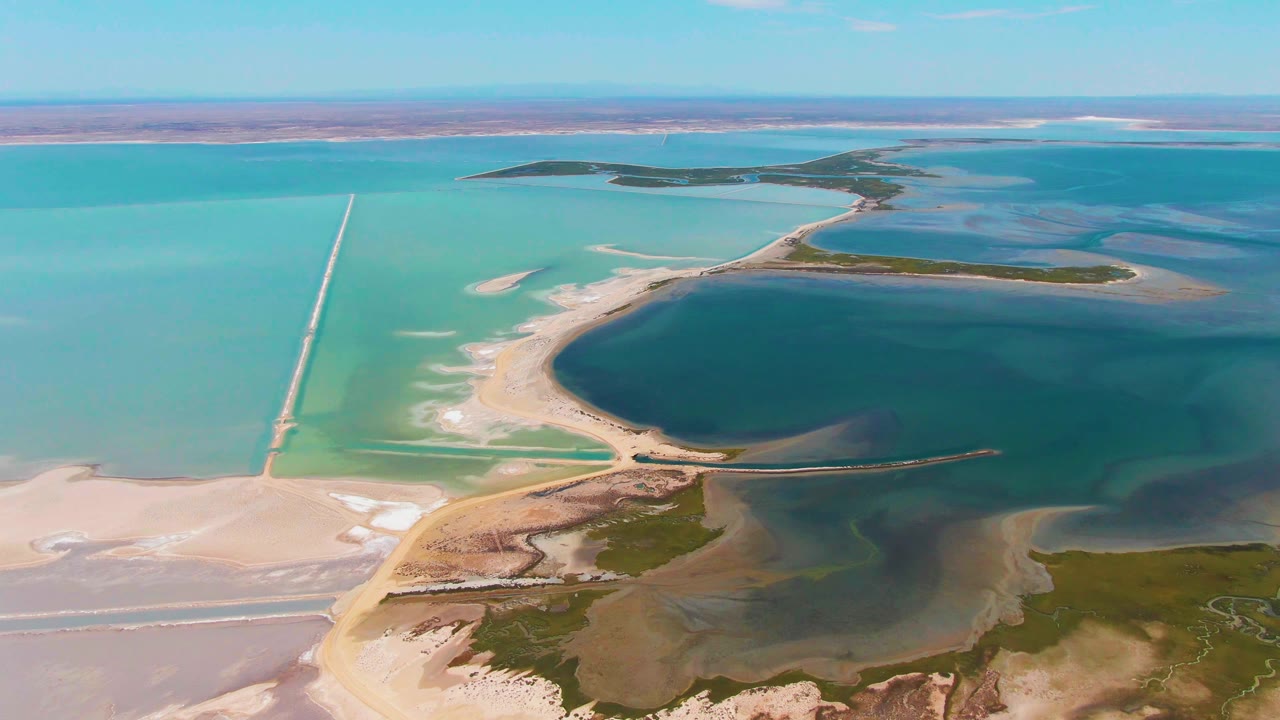 Second-frame aerial over Guerrero Negro ponds, showing the long pale causeway more centrally, blue-green water, salt-colored flats, low islands, and no visible people.
