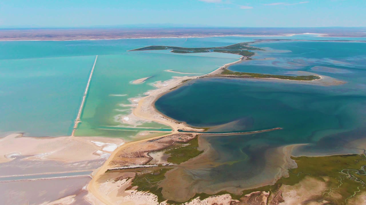 Aerial frame from Guerrero Negro with the dark evaporation basin on the right, turquoise pond water to the left, pale salt flats below, narrow causeways, and no visible people.