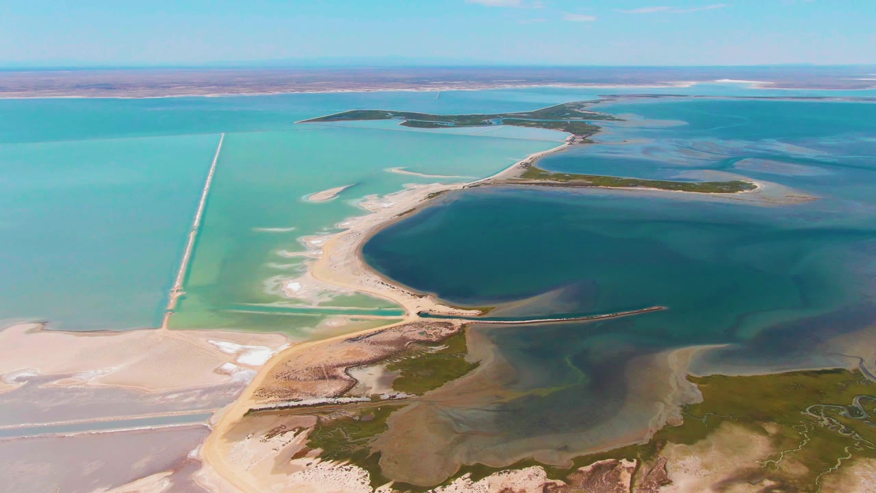 Mid-clip aerial over Guerrero Negro evaporation ponds, with salt flats in the lower left, green vegetation at the lower right, crossing causeways, layered blue water, and no visible people.