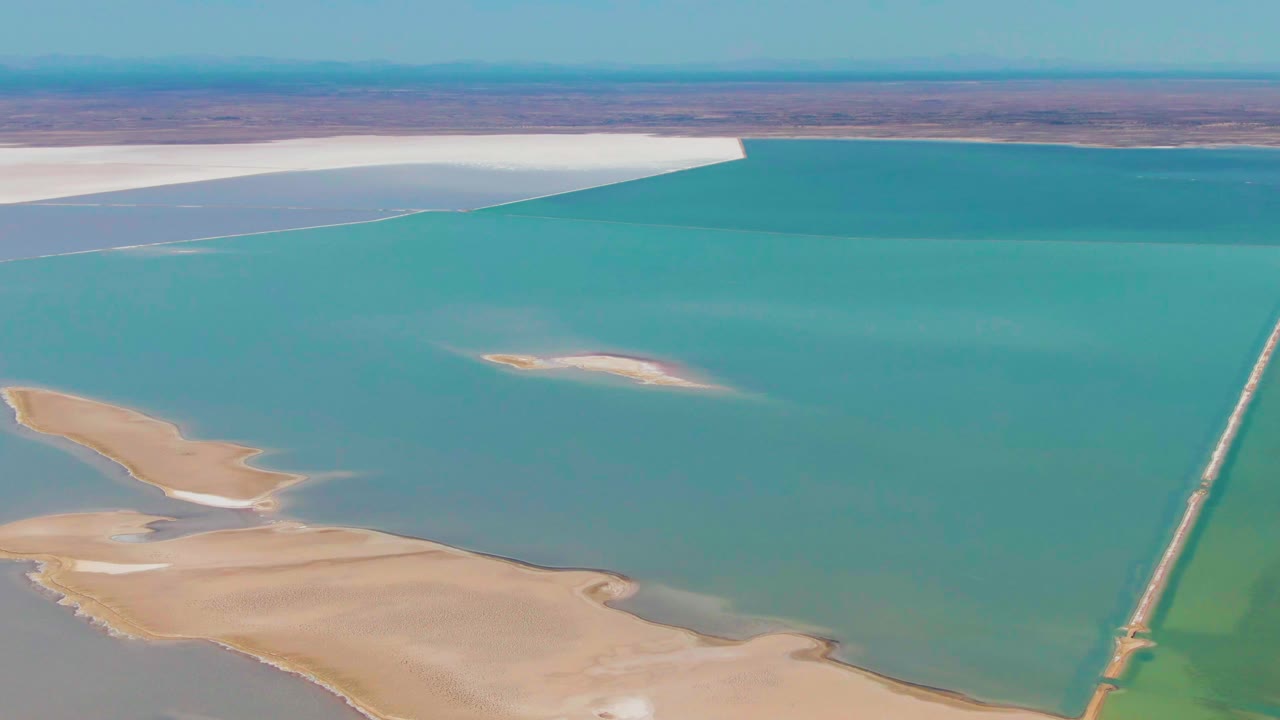 Aerial view in Guerrero Negro, Baja California Sur, with turquoise water filling most of the frame, sandbars lower left, a small pale island near center, the causeway on the right, and no visible people.