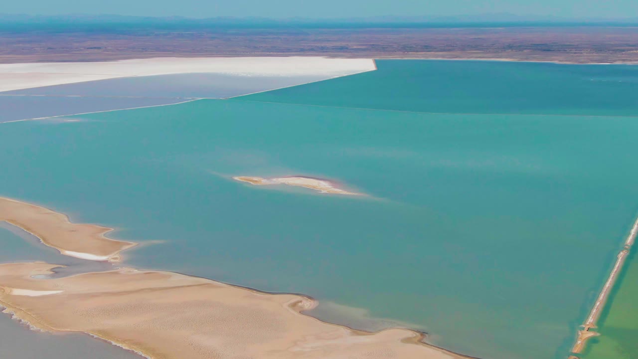 Aerial frame from Guerrero Negro, Baja California Sur, showing the turquoise salt lake, beige shoreline and sandbar shapes at lower left, a pale island in midwater, white flats across the top, and no visible people.