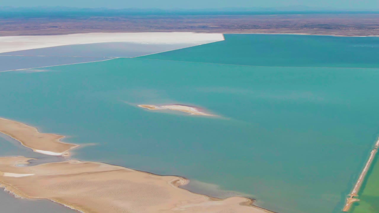 Mid-clip aerial frame over Guerrero Negro, Baja California Sur, with open turquoise water dominating the image, the causeway partly cropped at right, beige land low in frame, white flats at the far shore, and no visible people.