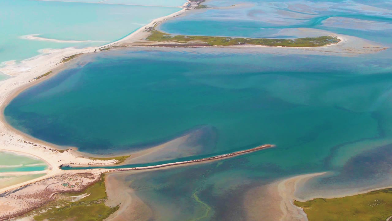 Aerial frame from Guerrero Negro, Baja California Sur centered on broad teal lagoon water, pale sand flats, a straight breakwater, and low green islands with no visible people.